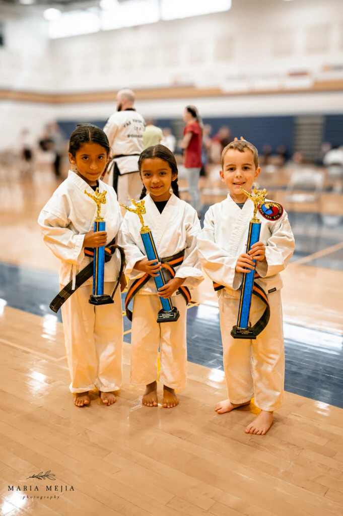 Three Champion Martial Arts students holding trophies in a gymnasium