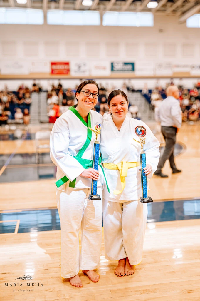 Two adult Champion Martial Arts students holding tournament trophies in a gym